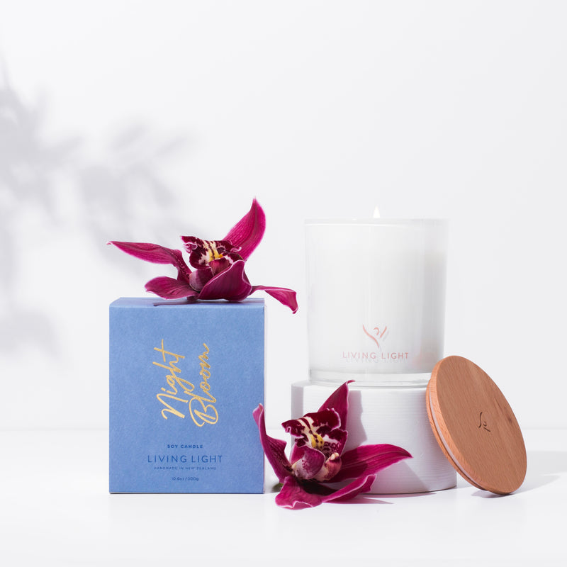 Living Light Night Bloom soy candle in white glass with wooden lid, displayed beside its blue packaging and purple orchids. Handcrafted in New Zealand.