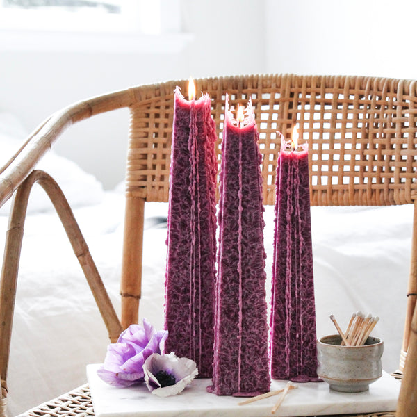 Lit Icicle Candles by Living Light NZ in plum tone, showcasing crystalline wax texture and sculptural drip effect, styled on a rattan chair with florals.