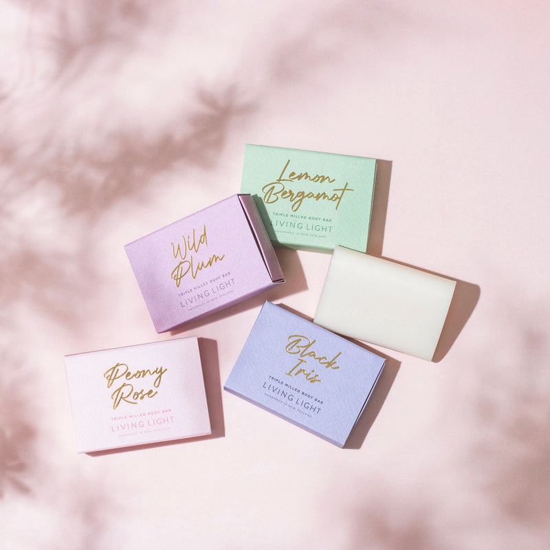 living light body bars in scents, Wild plum,Lemon bergamot, peony rose and Black iris on pink background with shadow of trees on left hand side and right hand bottom corner 